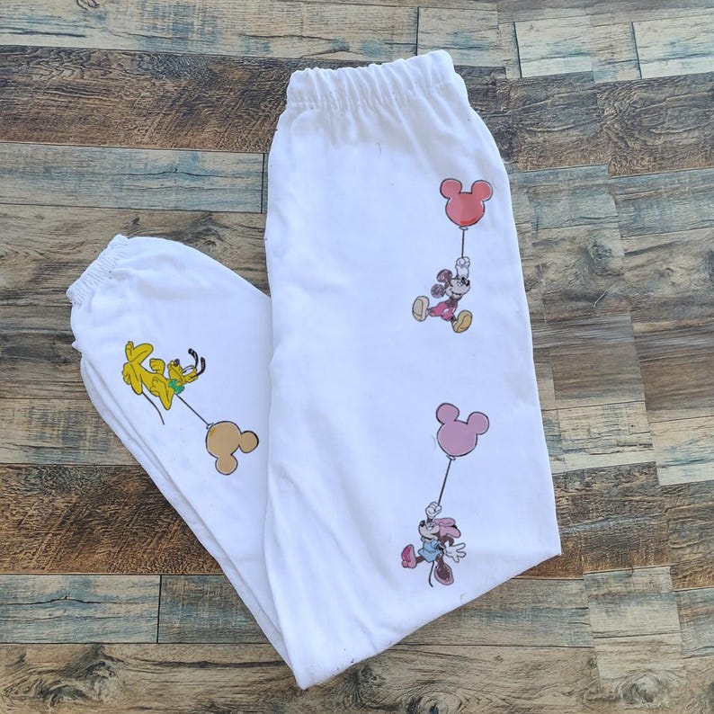 Pode incluir: Cal&ccedil;as de moletom brancas com personagens de desenhos animados segurando bal&otilde;es. As cal&ccedil;as t&ecirc;m c&oacute;s e punhos el&aacute;sticos. Os personagens de desenhos animados incluem Mickey Mouse, Minnie Mouse e Pluto. Os bal&otilde;es s&atilde;o rosa e bege.