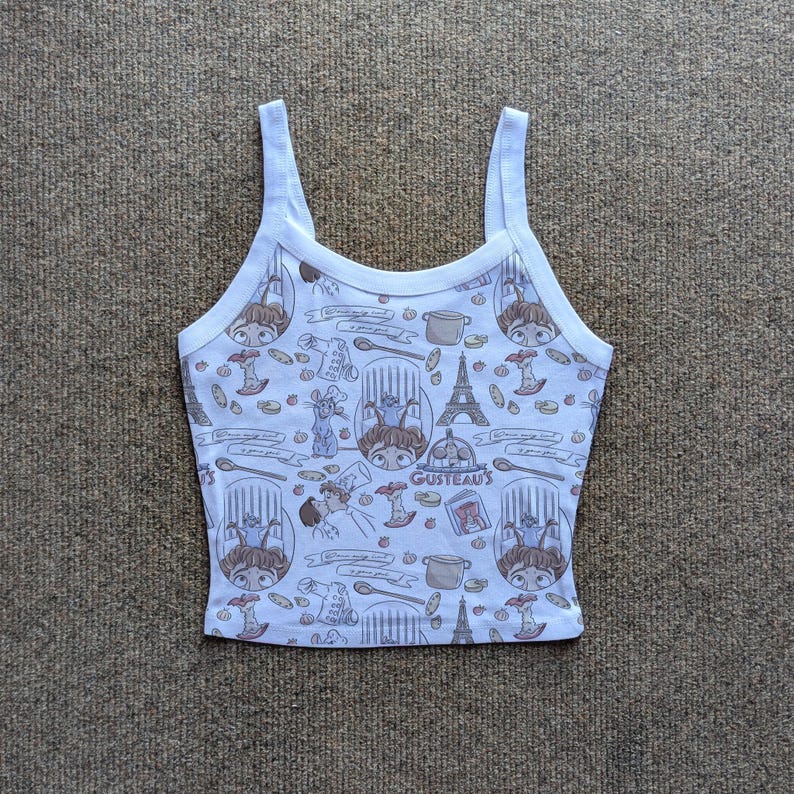 May include: White tank top with a repeating pattern of cartoon characters, cooking utensils, and the Eiffel Tower. The text "GUSTEAU'S" is visible on the front. The top has thin straps and a scoop neckline.