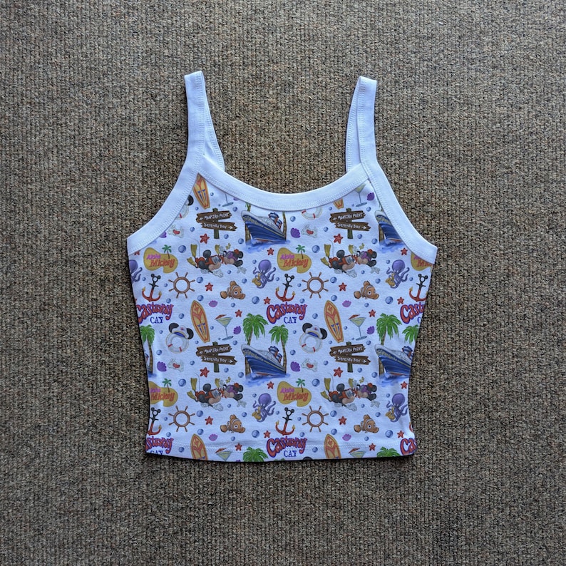 May include: White tank top with a colorful all-over print featuring cartoon characters, cruise ships, anchors, surfboards, and tropical elements. The top has a white trim and straps. The word "CAT" is visible on the print.