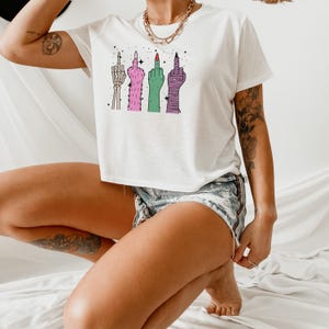 May include: White t-shirt featuring a graphic of four stylized middle fingers in various designs and colors. The fingers are depicted as a skeleton hand, a furry pink hand, a green hand, and a wrapped purple hand. The shirt is paired with denim shorts.