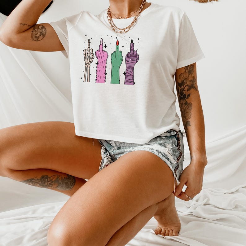 May include: White t-shirt featuring a graphic of four stylized middle fingers in various designs and colors. The fingers are depicted as a skeleton hand, a furry pink hand, a green hand, and a wrapped purple hand. The shirt is paired with denim shorts.