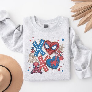 Puede incluir: Sudadera gris con un gr&aacute;fico de Spiderman rojo y azul. El gr&aacute;fico presenta la cara de Spiderman, un coraz&oacute;n y las letras XOXO.