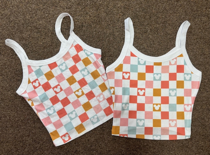 May include: Two white tank tops with a checkered pattern in pink, blue, and orange with a white Mickey Mouse silhouette repeated throughout the pattern.