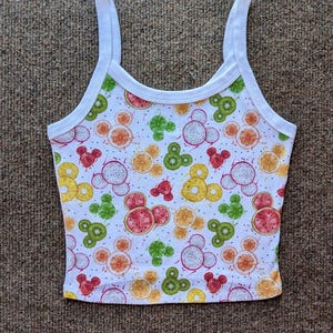 May include: White tank top with a fruit print featuring Mickey Mouse silhouettes. The fruit includes kiwi, watermelon, pineapple, and dragon fruit.