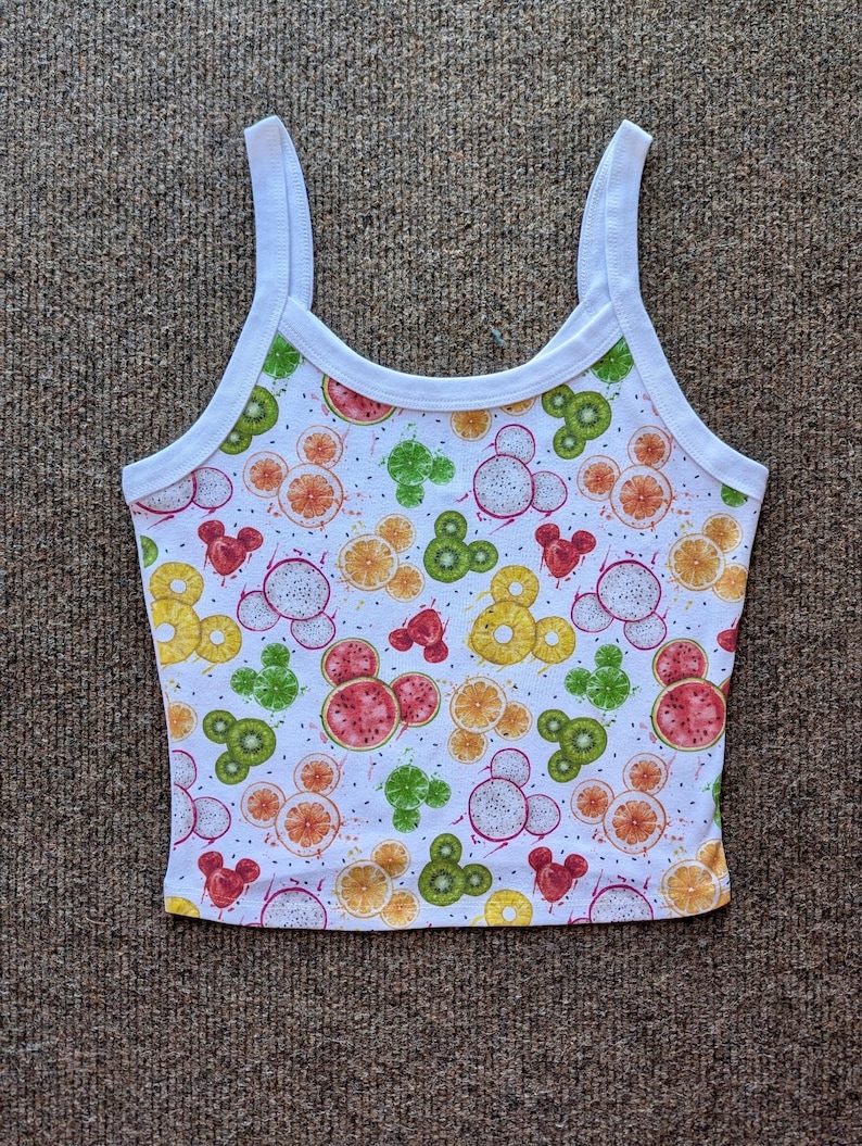 May include: White tank top with a fruit print featuring Mickey Mouse silhouettes. The fruit includes kiwi, watermelon, pineapple, and dragon fruit.