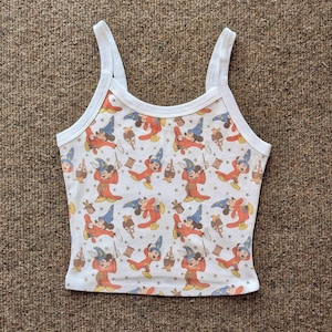 May include: White tank top with a repeating pattern of a cartoon character in a wizard costume. The character is wearing a blue hat and red robe. The tank top has white straps and trim.