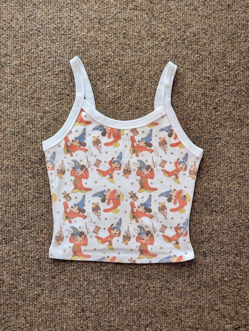 May include: White tank top with a repeating pattern of a cartoon character in a wizard costume. The character is wearing a blue hat and red robe. The tank top has white straps and trim.