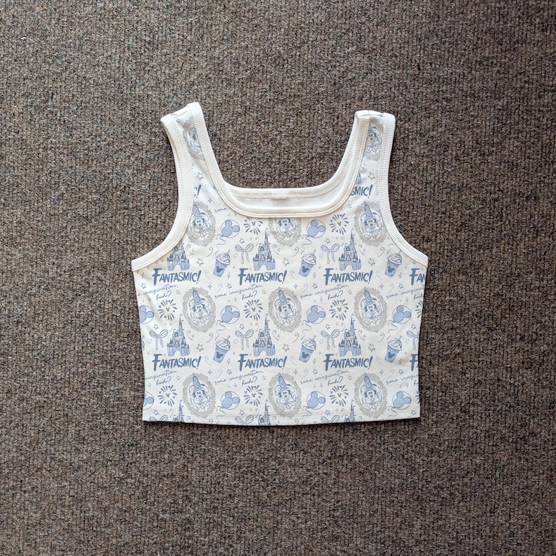 May include: White tank top with a repeating pattern of blue and gray Disney-themed graphics, including castles, Mickey Mouse, and the word "Fantasmic!". The top has a square neckline and wide straps.