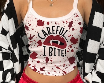 May include: White tank top with a red splatter design and the text "CAREFUL I BITE!" in black. The top has a scoop neck and thin straps. The model is wearing red satin shorts and a black and white checkered cardigan.