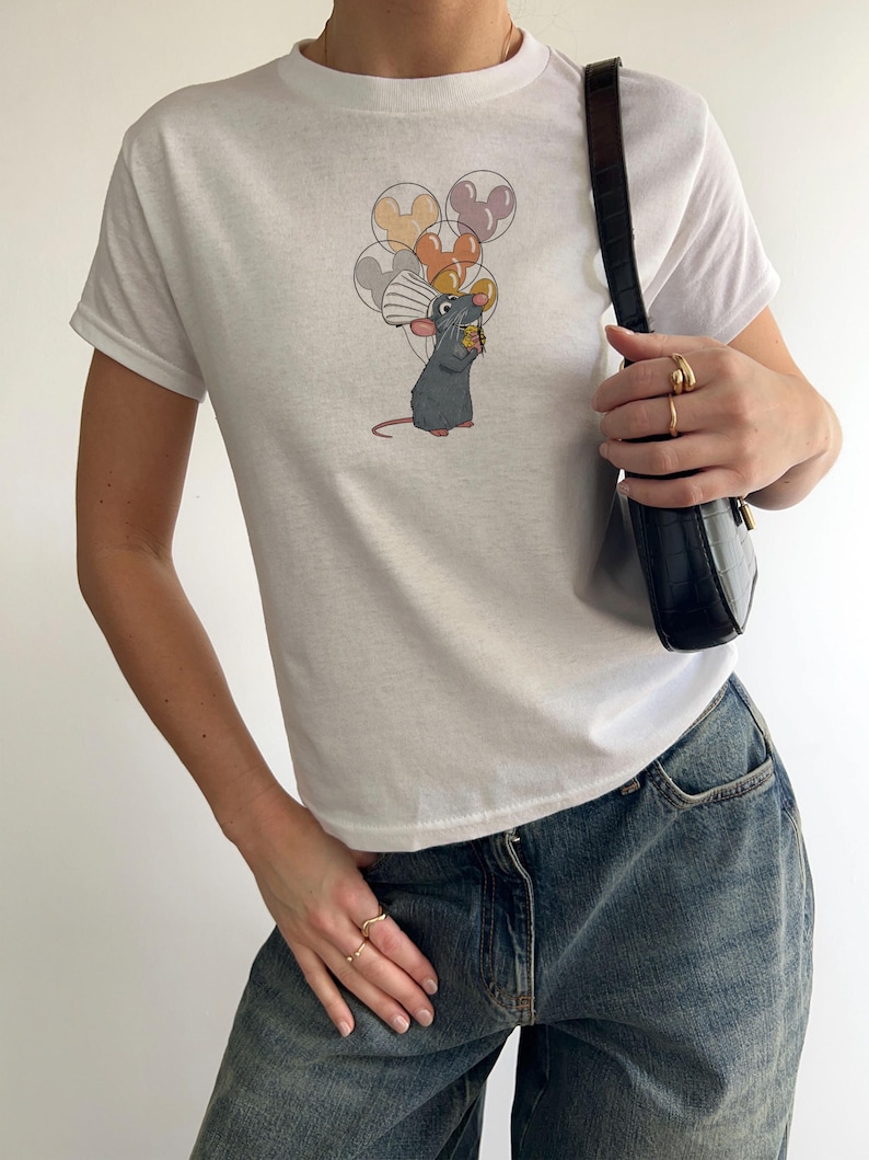 May include: A white t-shirt with a graphic of a rat holding a piece of cheese and a bunch of balloons with Mickey Mouse ears on them.