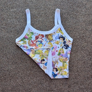 Puede incluir: Una camiseta sin mangas blanca con un estampado colorido con varias princesas de Disney. La camiseta tiene ribetes y tirantes blancos. El estampado incluye personajes como Ariel, Blancanieves y Bella, sobre un fondo claro.