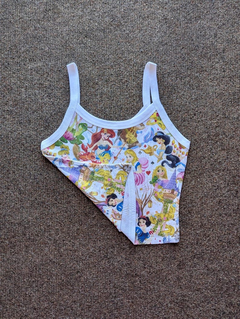 Puede incluir: Una camiseta sin mangas blanca con un estampado colorido con varias princesas de Disney. La camiseta tiene ribetes y tirantes blancos. El estampado incluye personajes como Ariel, Blancanieves y Bella, sobre un fondo claro.