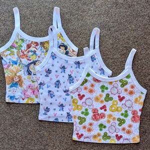 May include: Three white tank tops with different colorful cartoon prints. The first tank top has a print of Disney princesses, the second tank top has a print of Stitch from Lilo & Stitch, and the third tank top has a print of Mickey Mouse heads with fruit slices.