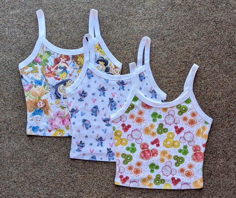 May include: Three white tank tops with different colorful cartoon prints. The first tank top has a print of Disney princesses, the second tank top has a print of Stitch from Lilo & Stitch, and the third tank top has a print of Mickey Mouse heads with fruit slices.