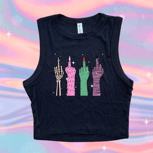 May include: Black sleeveless crop top featuring a graphic of four stylized hands giving the middle finger. The hands are depicted as a skeleton, pink fur, green skin, and wrapped in bandages. The nails are painted red and black.