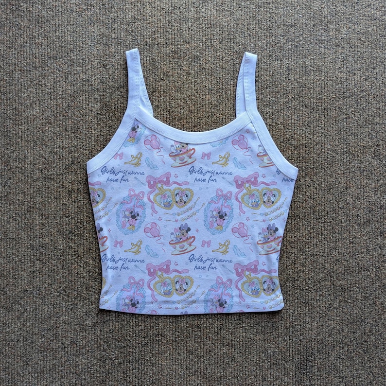 May include: White tank top with a repeating pattern of cartoon characters, teacups, bows, and the text "Girls just wanna have fun." The top has a scoop neckline and thin straps. The overall color scheme is pastel pink, yellow, and blue.