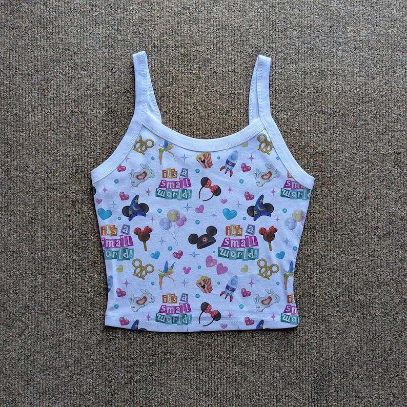 May include: White tank top with a colorful "It's a Small World" print. The design includes Mickey Mouse ears, hearts, rockets, and other whimsical elements. The top has thin straps and a rounded neckline.