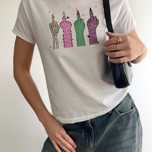 May include: White t-shirt featuring a graphic of four stylized hands giving the middle finger. The hands are depicted as a skeleton, furry, green, and wrapped in bandages. The shirt is paired with blue jeans.