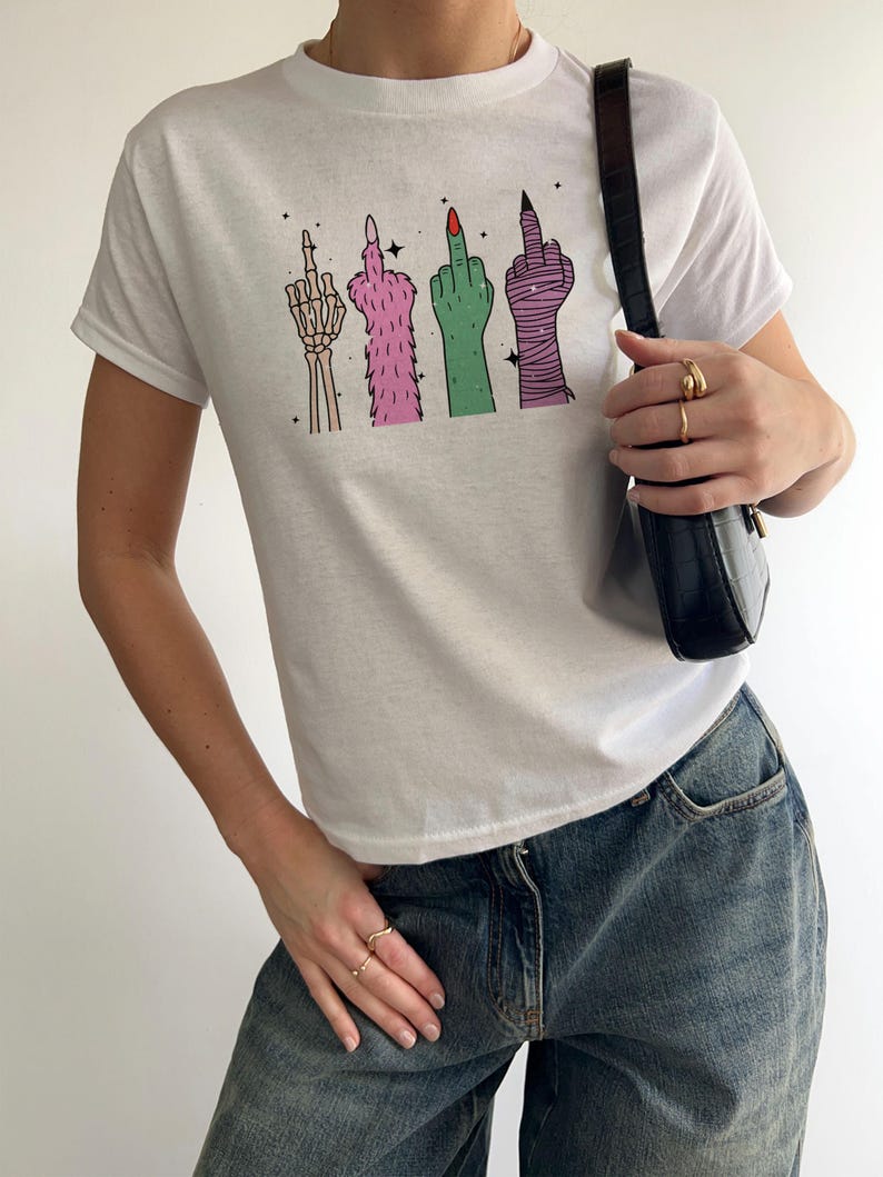 May include: White t-shirt featuring a graphic of four stylized hands giving the middle finger. The hands are depicted as a skeleton, furry, green, and wrapped in bandages. The shirt is paired with blue jeans.