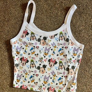 Puede incluir: Top corto blanco con un estampado repetido de Mickey Mouse, Minnie Mouse, Donald Duck, Daisy Duck y Goofy. La camiseta sin mangas tiene tirantes finos blancos y un ribete blanco alrededor del cuello y las sisas.