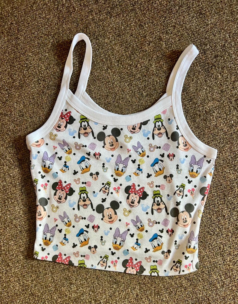 Puede incluir: Top corto blanco con un estampado repetido de Mickey Mouse, Minnie Mouse, Donald Duck, Daisy Duck y Goofy. La camiseta sin mangas tiene tirantes finos blancos y un ribete blanco alrededor del cuello y las sisas.