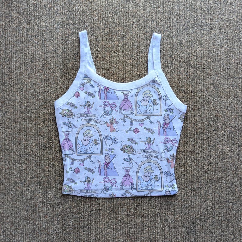 May include: White tank top with a repeating pattern of Cinderella-themed illustrations. The pattern includes Cinderella in a frame, dresses, mice, and text that reads "A dream is a wish your heart makes".