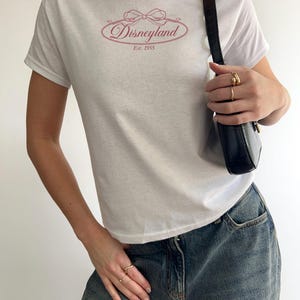 May include: White t-shirt featuring a pink Disneyland logo with a bow and the text "Disneyland Est. 1955". The shirt is paired with blue denim jeans and a black shoulder bag. The person is wearing gold rings.