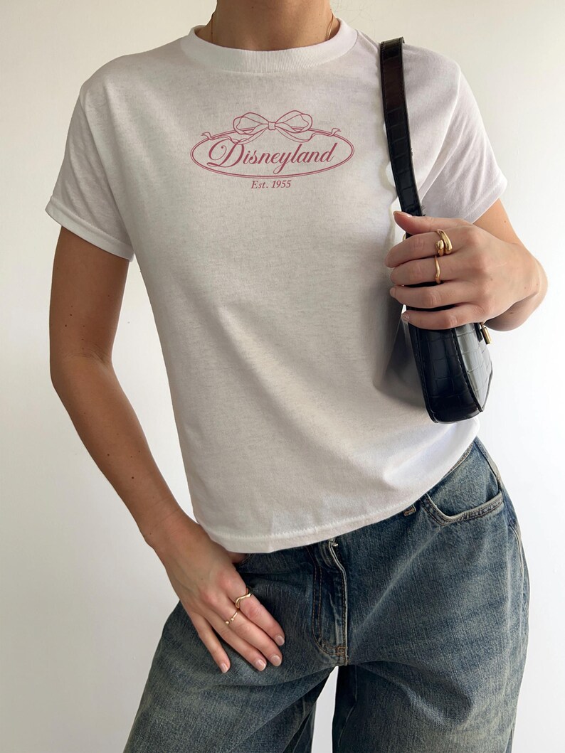 May include: White t-shirt featuring a pink Disneyland logo with a bow and the text "Disneyland Est. 1955". The shirt is paired with blue denim jeans and a black shoulder bag. The person is wearing gold rings.