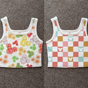 May include: Two white crop tops with a square neckline. One top features a fruit pattern with Mickey Mouse shapes, including watermelon, kiwi, and orange slices. The other top has a checkered pattern in pink, orange, and blue with Mickey Mouse silhouettes.