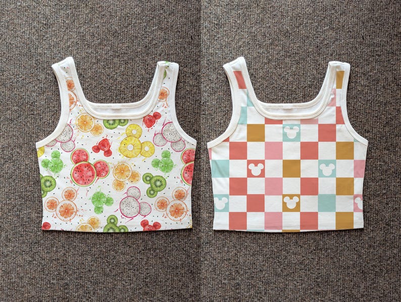 May include: Two white crop tops with a square neckline. One top features a fruit pattern with Mickey Mouse shapes, including watermelon, kiwi, and orange slices. The other top has a checkered pattern in pink, orange, and blue with Mickey Mouse silhouettes.