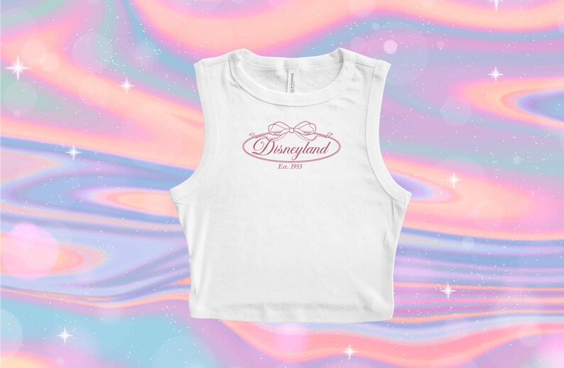 May include: White sleeveless crop top with a pink Disneyland logo. The logo features the word "Disneyland" in a script font, with a bow above and "Est. 1955" below. The background is a colorful, swirling pattern of pink, blue, and purple.