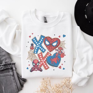 Puede incluir: Sudadera blanca con un dise&ntilde;o gr&aacute;fico que presenta un coraz&oacute;n rojo con una m&aacute;scara de Spider-Man, un dise&ntilde;o XOXO rojo y azul y un personaje de Spider-Man.