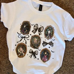 May include: White t-shirt with a print of six Disney villains in gold frames with black bows and bats. The villains are Maleficent, Cruella de Vil, Ursula, Evil Queen, Queen of Hearts, and Hades.
