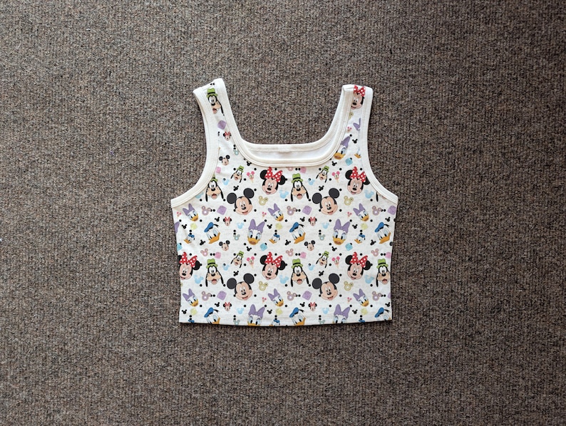 Puede incluir: Top corto blanco con un estampado colorido con Mickey Mouse, Minnie Mouse, Donald Duck y Daisy Duck. El top sin mangas tiene cuello redondo y tirantes anchos. La tela es un material suave y ligero.