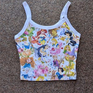 May include: White tank top with a colorful print of various Disney princesses and characters. The print includes Ariel, Cinderella, Snow White, and Alice in Wonderland.
