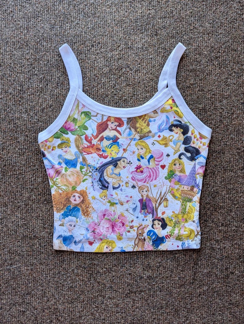 May include: White tank top with a colorful print of various Disney princesses and characters. The print includes Ariel, Cinderella, Snow White, and Alice in Wonderland.