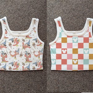 May include: Two white tank tops. One features a pattern of Mickey Mouse as a sorcerer in red and blue robes. The other has a checkerboard pattern in pink, orange, yellow, and teal, with Mickey Mouse silhouettes.