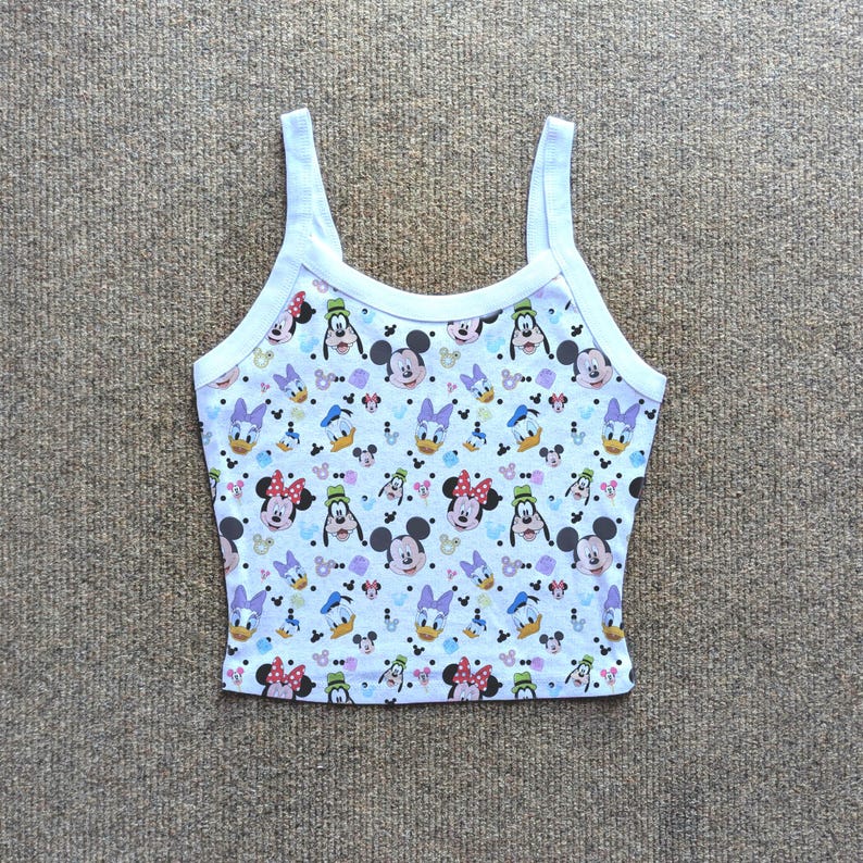Puede incluir: Camiseta sin mangas blanca con un estampado de personajes de Disney, incluyendo Mickey Mouse, Minnie Mouse, Donald Duck y Daisy Duck. La camiseta tiene ribetes y tirantes blancos. El fondo es de color azul claro.