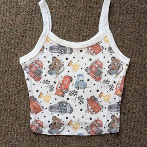 May include: White tank top with a colorful pattern of cartoon cars, bears, and stars. The top has thin white straps and a white trim around the neckline and armholes. The pattern includes red, blue, and brown vehicles.