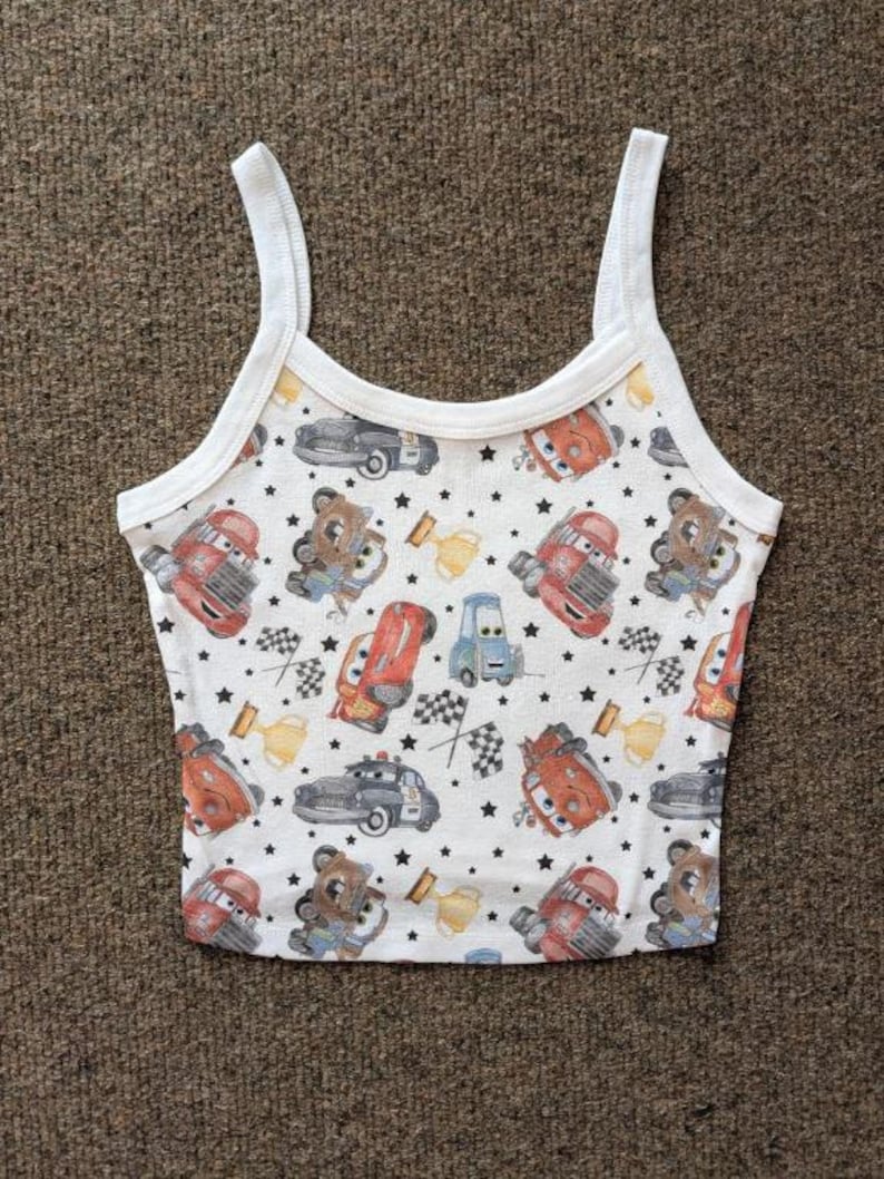May include: White tank top with a colorful pattern of cartoon cars, bears, and stars. The top has thin white straps and a white trim around the neckline and armholes. The pattern includes red, blue, and brown vehicles.