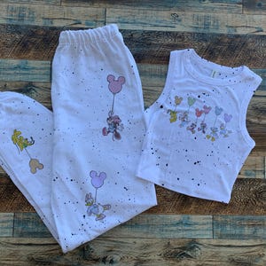 Pode incluir: Conjunto de pijama branco com top e cal&ccedil;as. O conjunto apresenta personagens de desenhos animados segurando bal&otilde;es. O conjunto &eacute; decorado com respingos de tinta preta. O top e as cal&ccedil;as s&atilde;o feitos de um material macio.