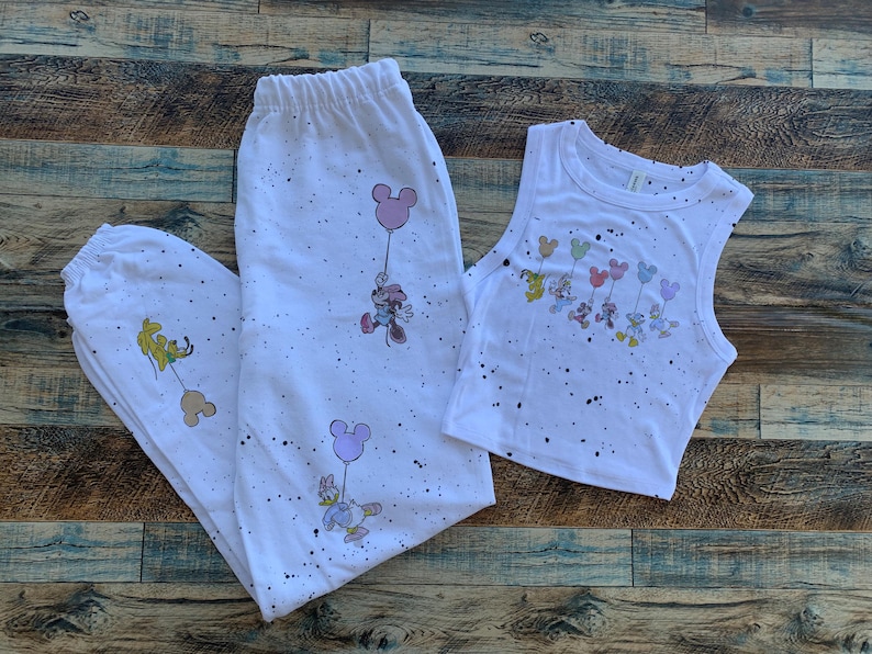 Pode incluir: Conjunto de pijama branco com top e cal&ccedil;as. O conjunto apresenta personagens de desenhos animados segurando bal&otilde;es. O conjunto &eacute; decorado com respingos de tinta preta. O top e as cal&ccedil;as s&atilde;o feitos de um material macio.