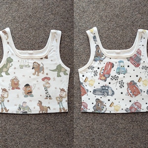 May include: Two white tank tops with a square neckline. One top features Toy Story characters like Woody and Buzz Lightyear. The other top showcases Cars characters, race flags, and stars. Both tops have a repeating pattern.