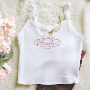 Może przedstawiać: Biały top na ramiączkach z napisem "Disneyland" w różowym kolorze, ze wstążką i napisem "Est. 1955". Złoty naszyjnik z zawieszką w kształcie motyla wisi na górze. Brązowe spodnie są widoczne na dole.