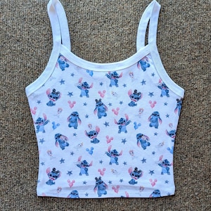May include: White tank top with a blue Stitch and Mickey Mouse print. The print features Stitch in various poses with Mickey Mouse ears and balloons. The background is white with white stars.