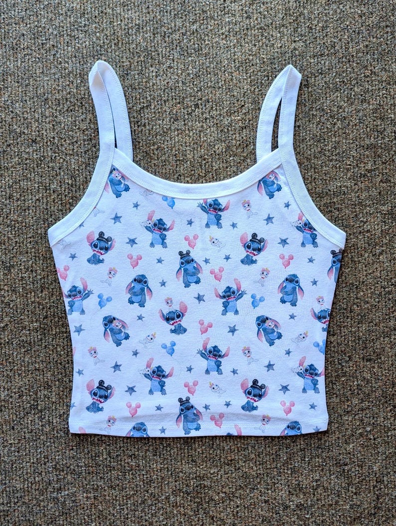 May include: White tank top with a blue Stitch and Mickey Mouse print. The print features Stitch in various poses with Mickey Mouse ears and balloons. The background is white with white stars.