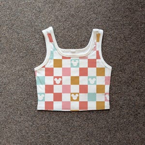 May include: White tank top with a checkerboard pattern in coral, pink, teal, and gold squares. Mickey Mouse head silhouettes are interspersed throughout the design. The top has a scoop neck and wide straps.