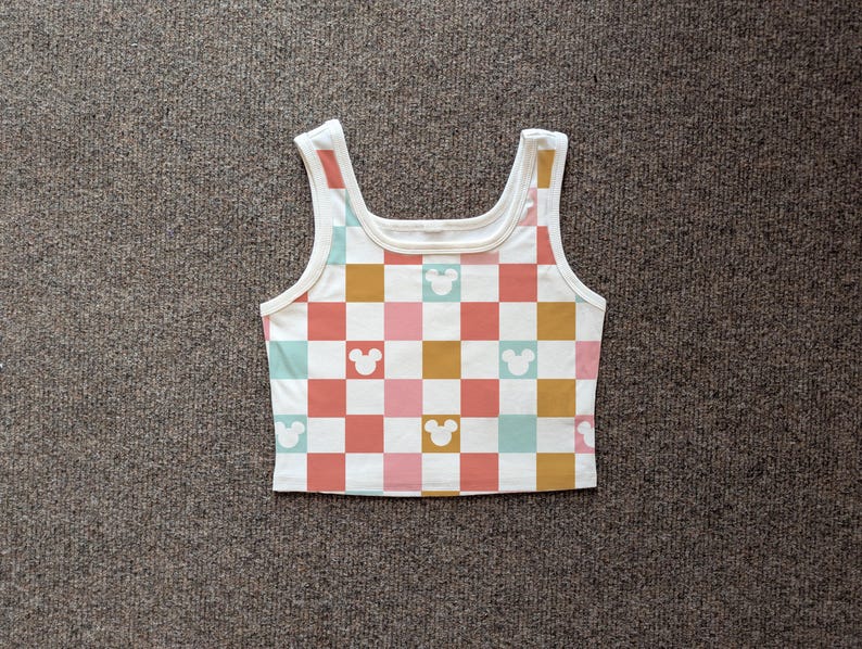 May include: White tank top with a checkerboard pattern in coral, pink, teal, and gold squares. Mickey Mouse head silhouettes are interspersed throughout the design. The top has a scoop neck and wide straps.
