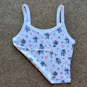 Puede incluir: Camiseta sin mangas blanca con un estampado repetido del personaje Stitch de Lilo & Stitch, estrellas y globos rosas con forma de Mickey Mouse. La camiseta sin mangas tiene tirantes finos y ribete blanco.