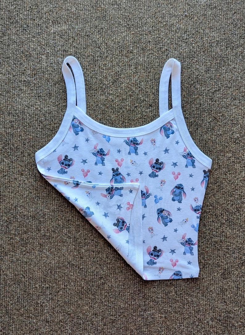 Puede incluir: Camiseta sin mangas blanca con un estampado repetido del personaje Stitch de Lilo & Stitch, estrellas y globos rosas con forma de Mickey Mouse. La camiseta sin mangas tiene tirantes finos y ribete blanco.